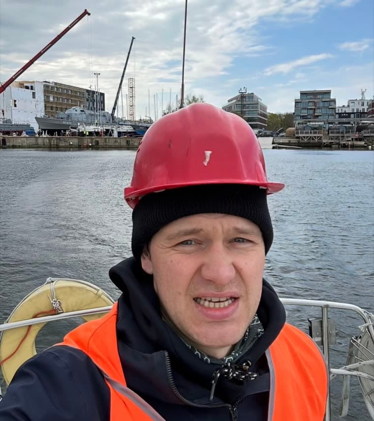 Ott Ilves in a red hard hat and an orange safety vest, with cranes behind him